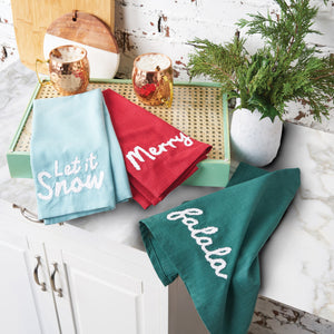 holiday towels with festive sayings in white on solid colored backgrounds layered on a kitchen counter