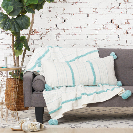 woven white pillow with light blue pom pom edges and tufting on a couch with a matching throw