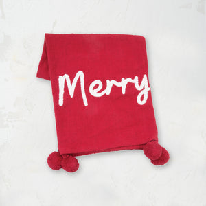 woven red throw with pom pom edges and the words merry in white